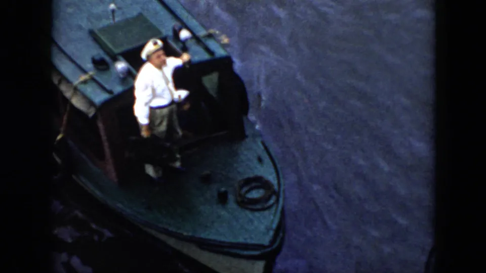 1956 Cuba frame showing the man in a white sailor-style uniform on the green motorboat, with rippled water and boat details visible.