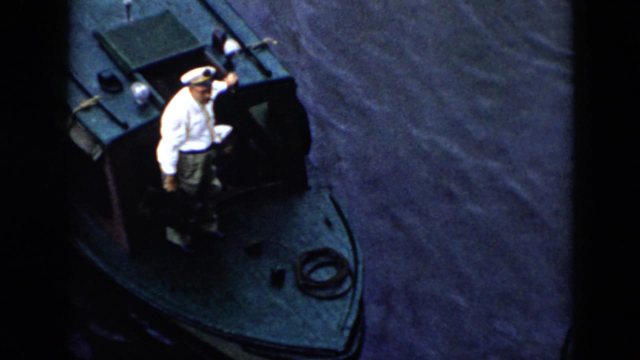 1956 Cuba home-movie frame showing a uniformed man in a white cap on the bow of a small dark boat, filmed from above.