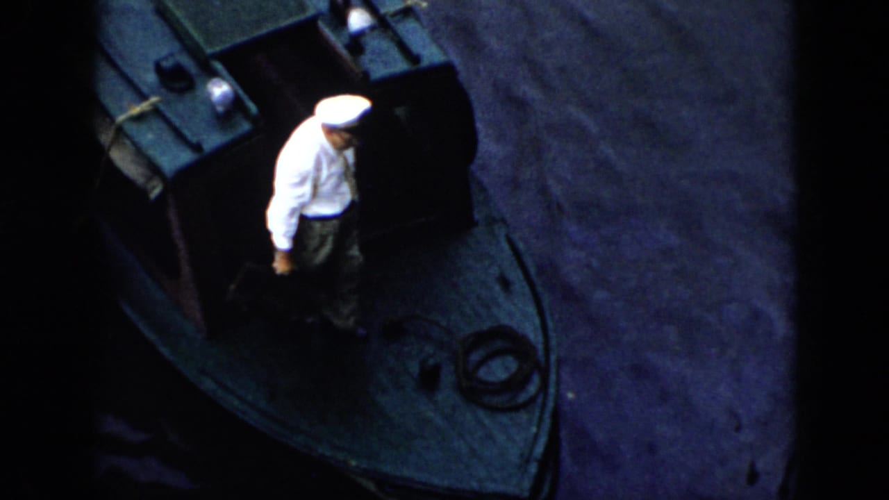 Man in a white uniform and cap walking on the small boat deck beside calm water.