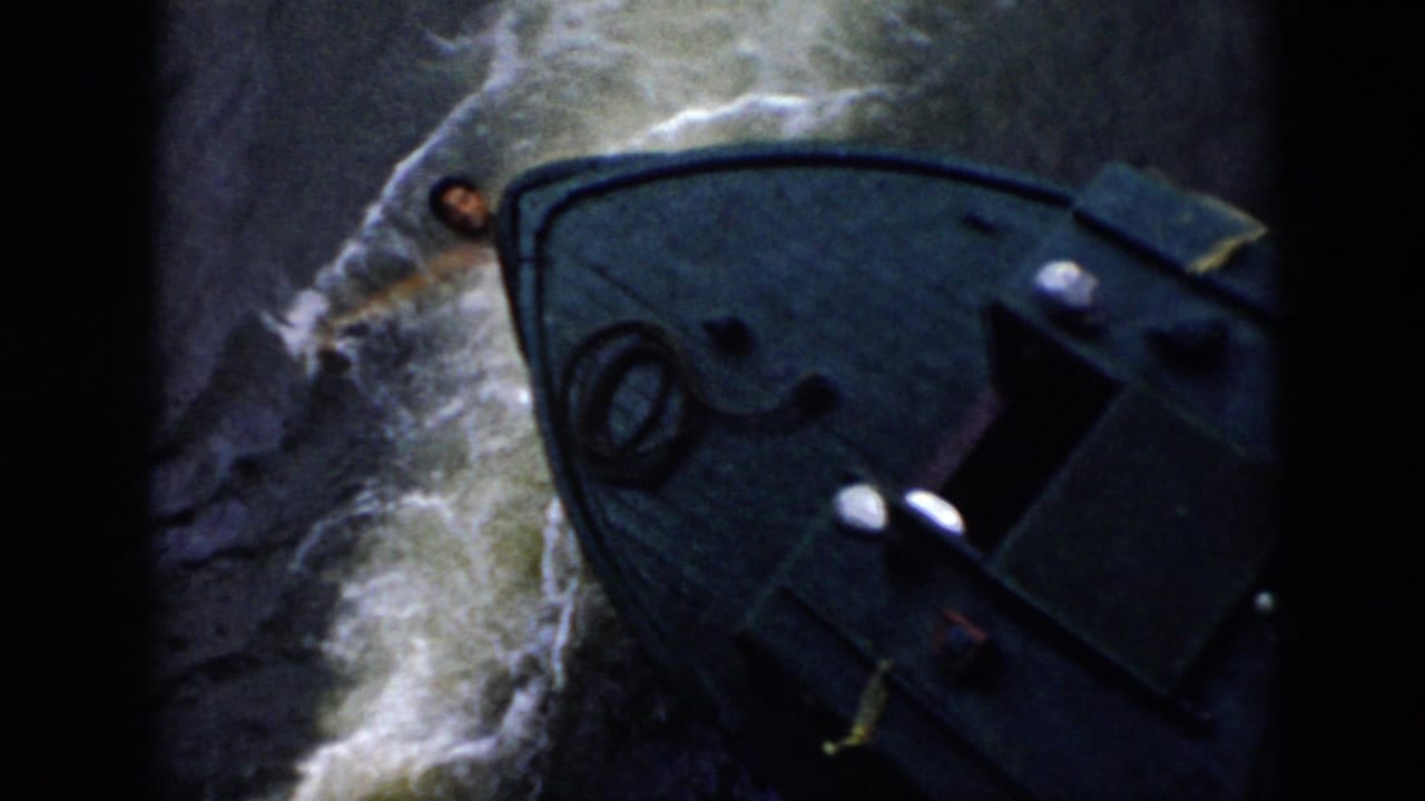 Boat bow moves past a man in the water with foamy waves along the hull.
