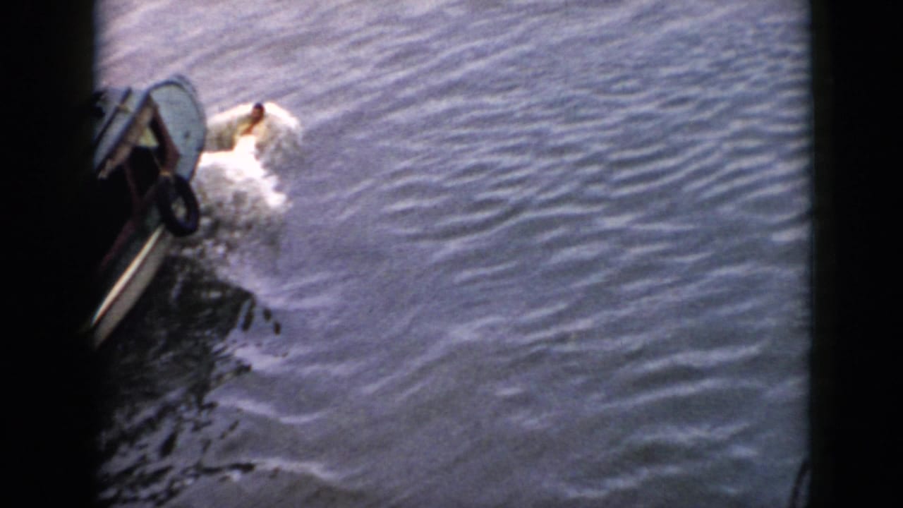 1956 Cuba home-movie frame with a swimmer raising an arm beside a small boat on choppy water.