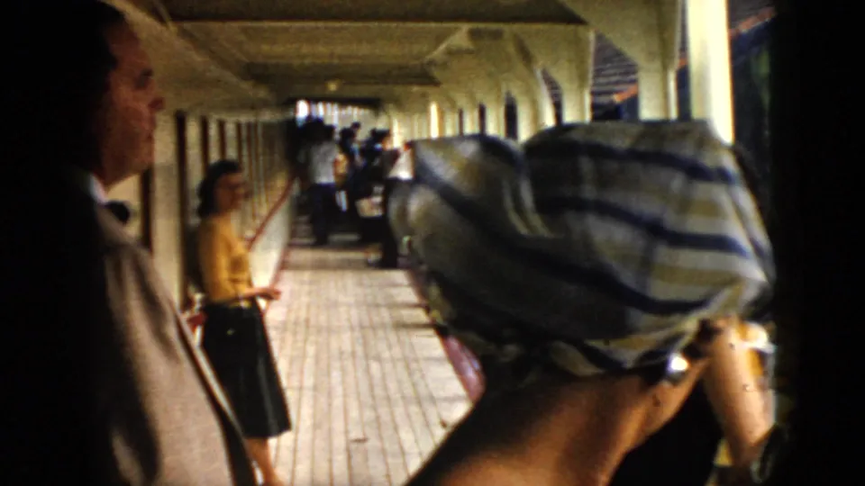 1956 Cuba home-movie frame of adults on a covered tiled walkway near railings, with a foreground striped head covering.