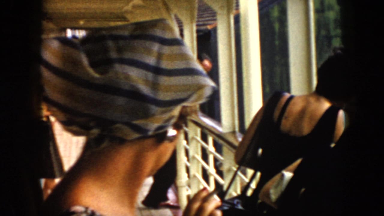 In 1956 Cuba home-movie footage, adults on a covered balcony lean and look outward, with a striped headscarf prominent at left.