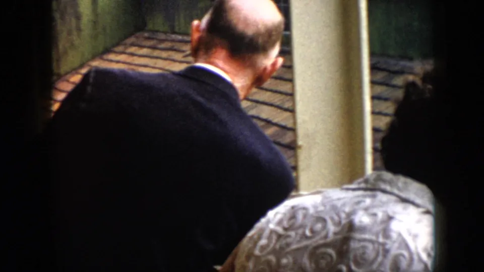 A man in a dark suit leans over the railing on a Cuban balcony, with roof tiles and columns visible ahead.