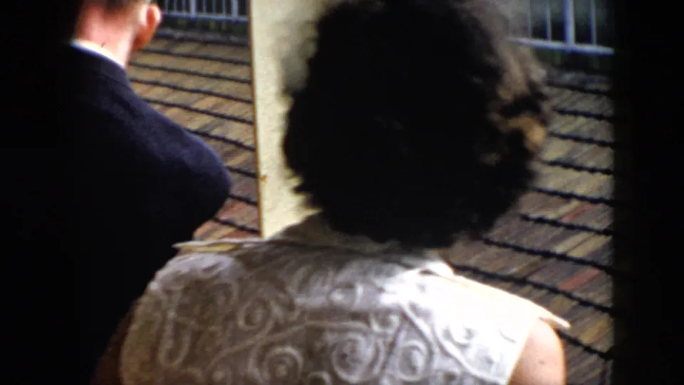 Two adults in 1956 Cuba stand with their backs to the camera on a covered balcony, watching an unseen subject beyond the railing.