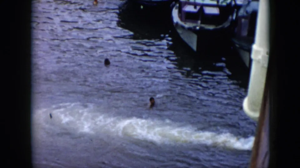 1956 Cuba home-movie view down to swimmers in the water, with small boats moored nearby and a wake crossing the frame.