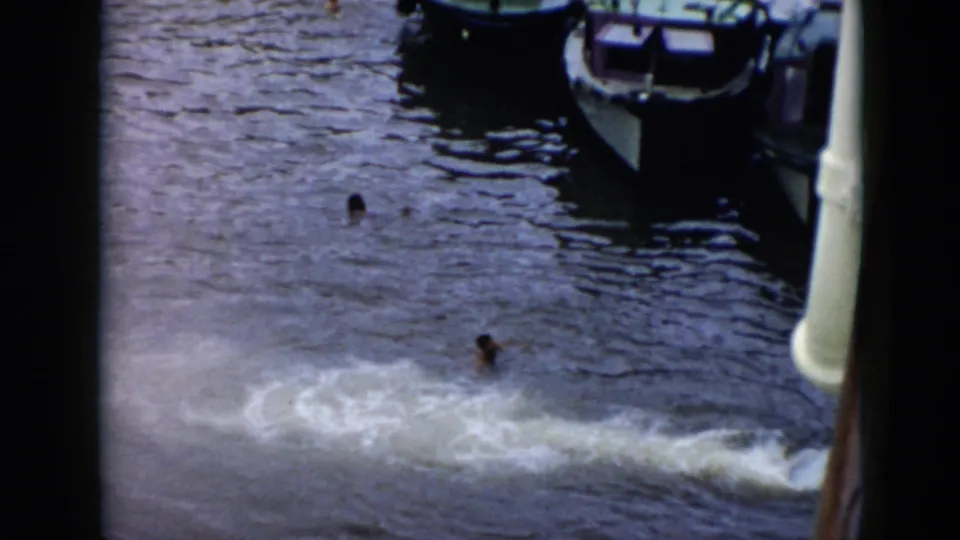 1956 Cuba frame looking down at swimmers in dark water near small boats, with foam or wake spreading across the foreground.