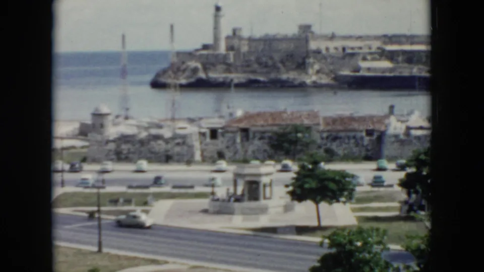1956 Cuba waterfront view with cars on a road, trees, harbor water, and a historic fortress-like structure beyond.
