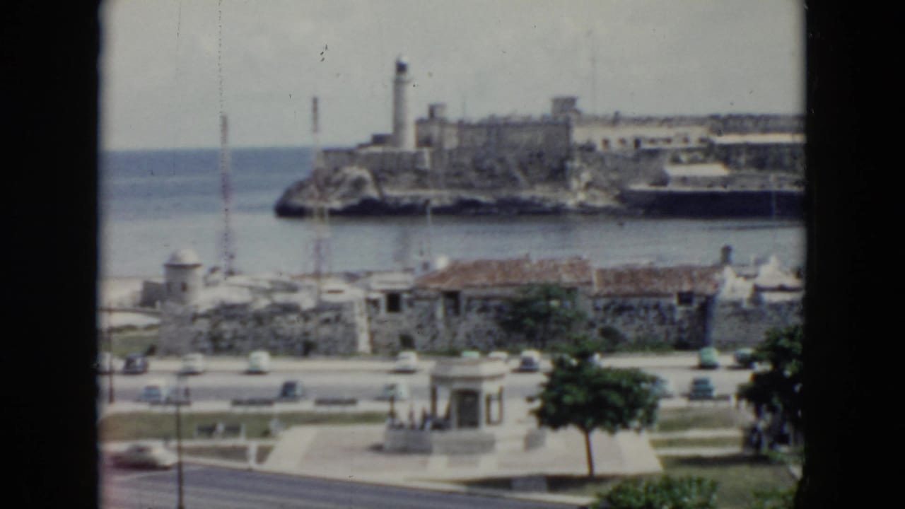 1956 Cuba waterfront seen through a window frame, with ocean, lighthouse, stone fortifications, road traffic, and small foreground buildings.