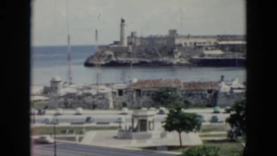 1956 Cuba waterfront seen through a window frame, with ocean, lighthouse, stone fortifications, road traffic, and small foreground buildings.