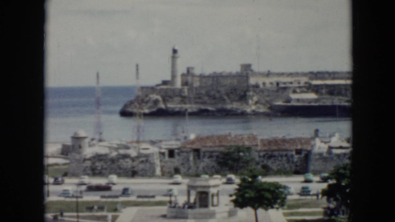 1956 Cuba coastal view with a lighthouse on rocky fortifications, ocean water, small buildings, cars, and a dark window edge.
