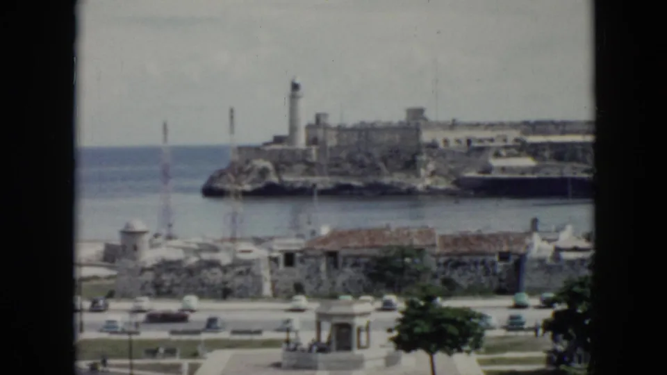 1956 Cuba coastal view with a lighthouse on rocky fortifications, ocean water, small buildings, cars, and a dark window edge.