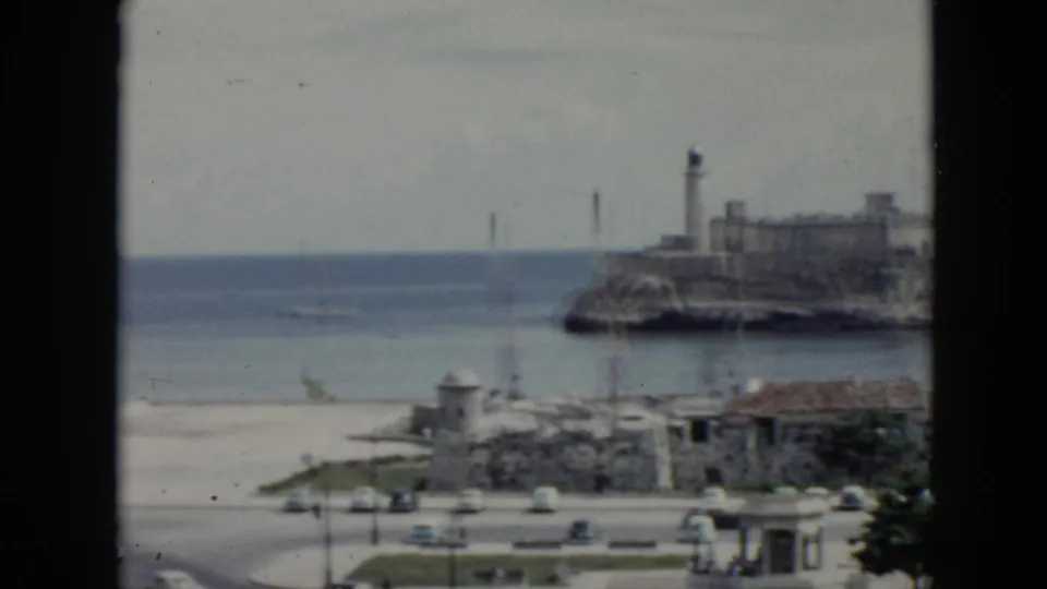 1956 Cuba waterfront in soft focus, showing ocean, a lighthouse, stone coastal buildings, red-tiled roofs, a road, and small cars.