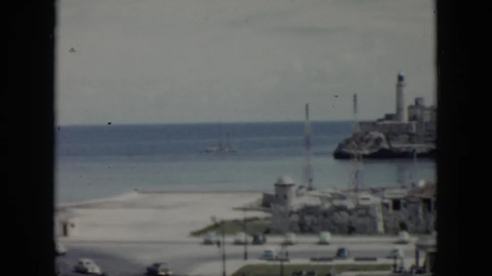 1956 Cuba coastal frame with the lighthouse partly at right, open sea, pale sky, road traffic, and low waterfront buildings.