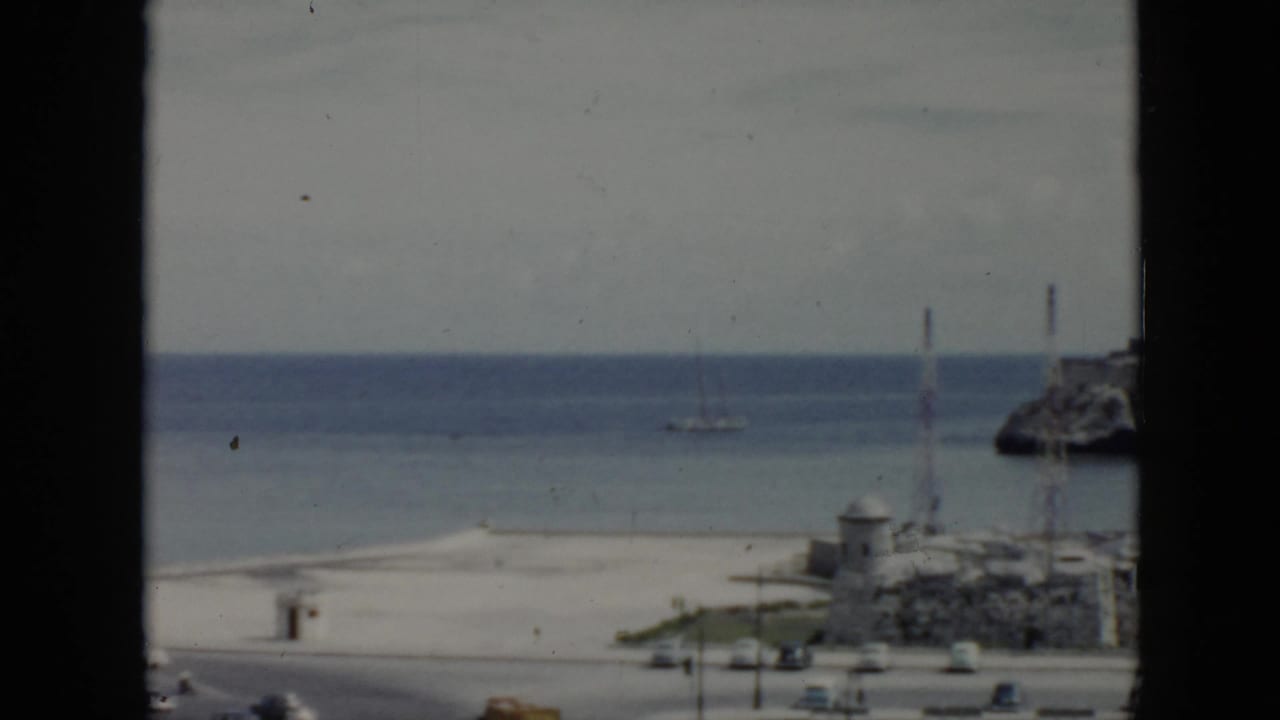 1956 Cuba 8mm frame showing a beach and calm ocean through a dark window frame, with a distant sailboat and road traffic below.