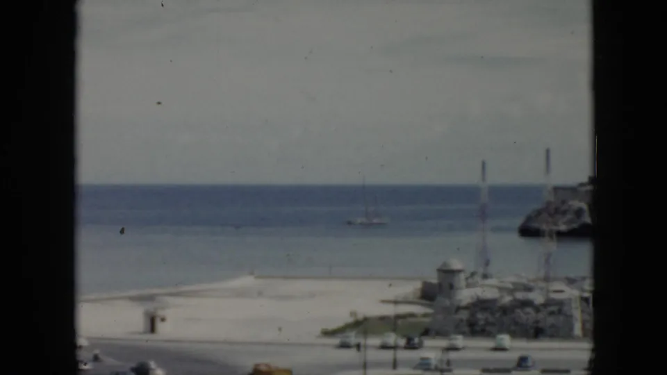 1956 Cuba 8mm frame showing a beach and calm ocean through a dark window frame, with a distant sailboat and road traffic below.
