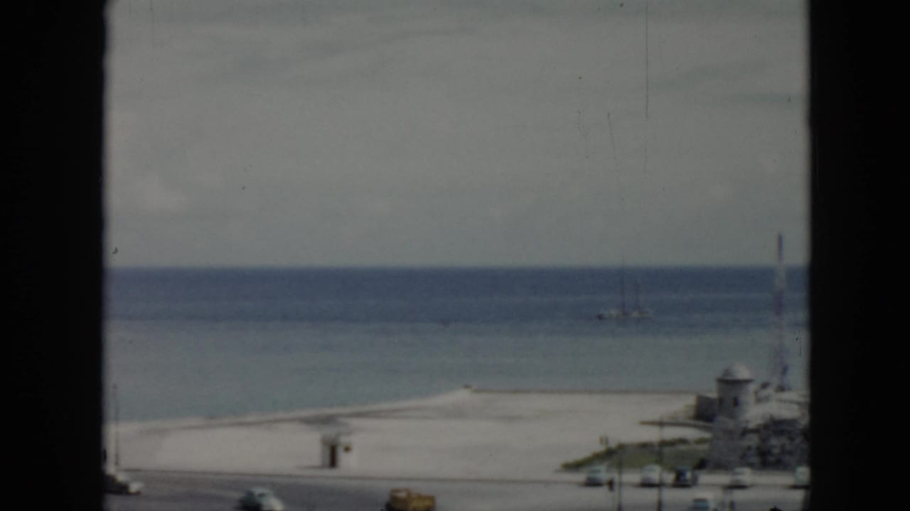 1956 Cuba 8mm frame of a quiet beach and ocean beyond a road, divided by a dark window frame with sailboats in the distance.