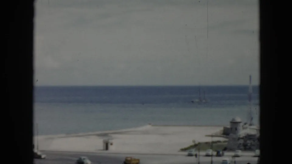 1956 Cuba 8mm frame of a quiet beach and ocean beyond a road, divided by a dark window frame with sailboats in the distance.