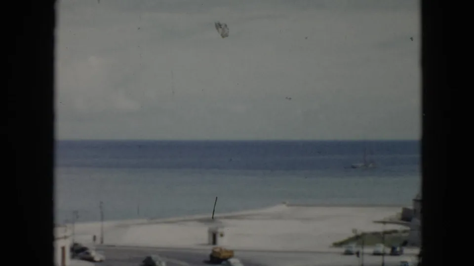 1956 Cuba 8mm frame showing a beach and ocean from an elevated window-framed viewpoint, with cars moving along the coastal road.