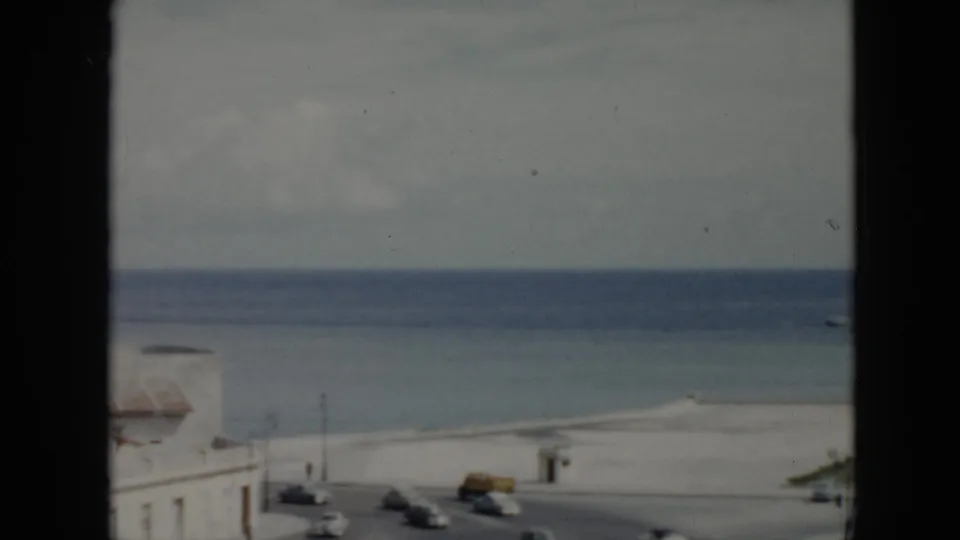 1956 Cuba 8mm frame with a window-framed view of a beachfront road, pale shoreline building, calm ocean, and distant boat.
