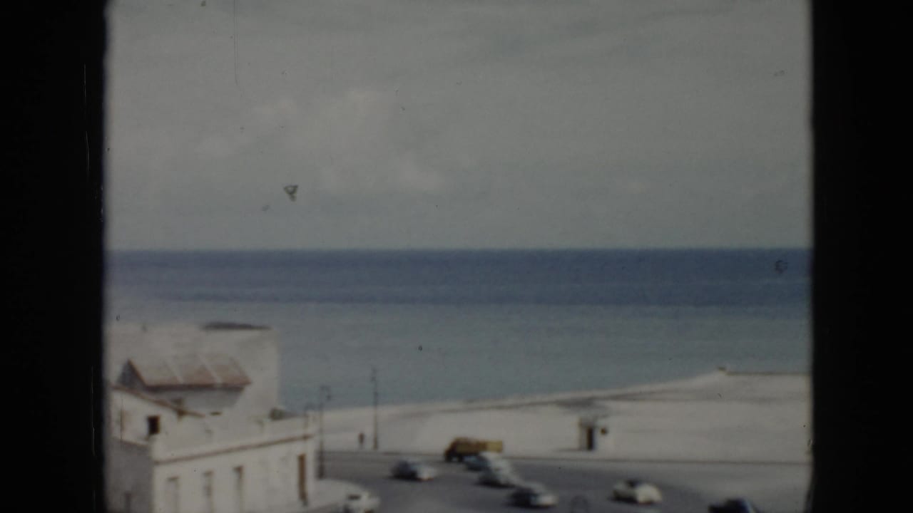 1956 Cuba waterfront view with white buildings, a curved road, moving cars, and the ocean beyond, partially blocked by a dark window frame.