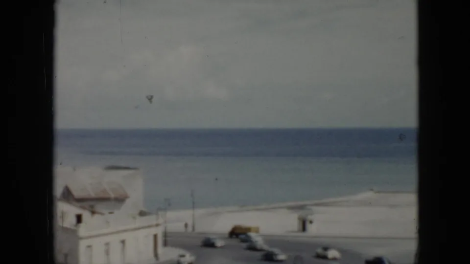 1956 Cuba waterfront view with white buildings, a curved road, moving cars, and the ocean beyond, partially blocked by a dark window frame.