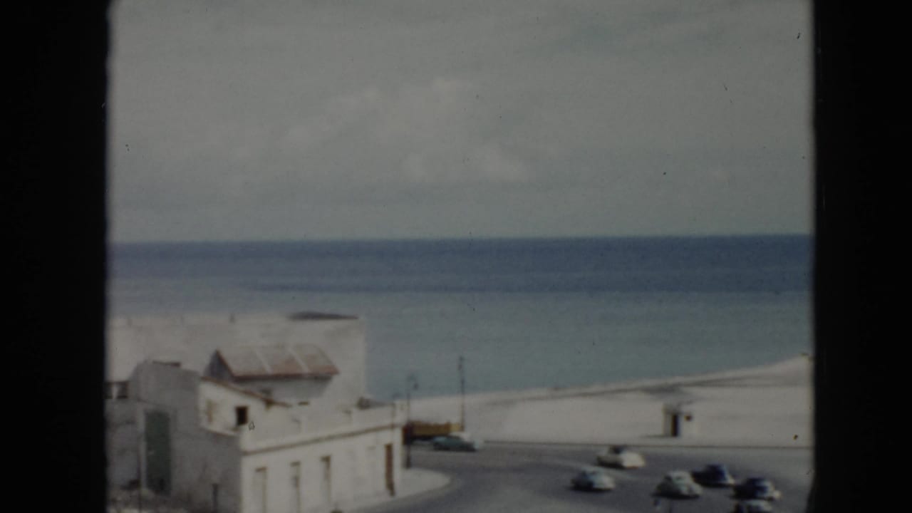 1956 Cuba coastal street scene with pale buildings, a seafront road, several cars, and the ocean visible behind them.