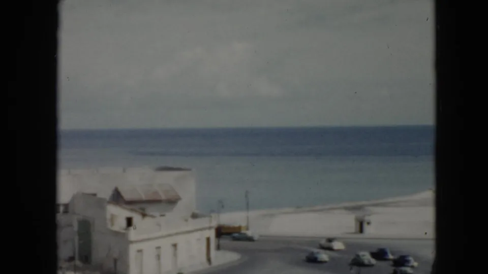 1956 Cuba coastal street scene with pale buildings, a seafront road, several cars, and the ocean visible behind them.