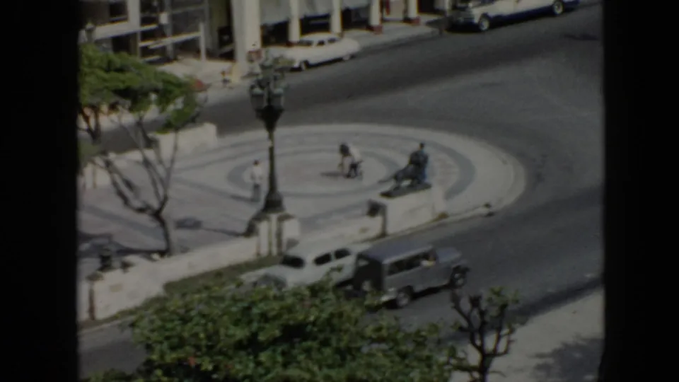 1956 Cuba urban waterfront view with a circular plaza, benches, trees, a station wagon, other cars, and small pedestrians near the road.