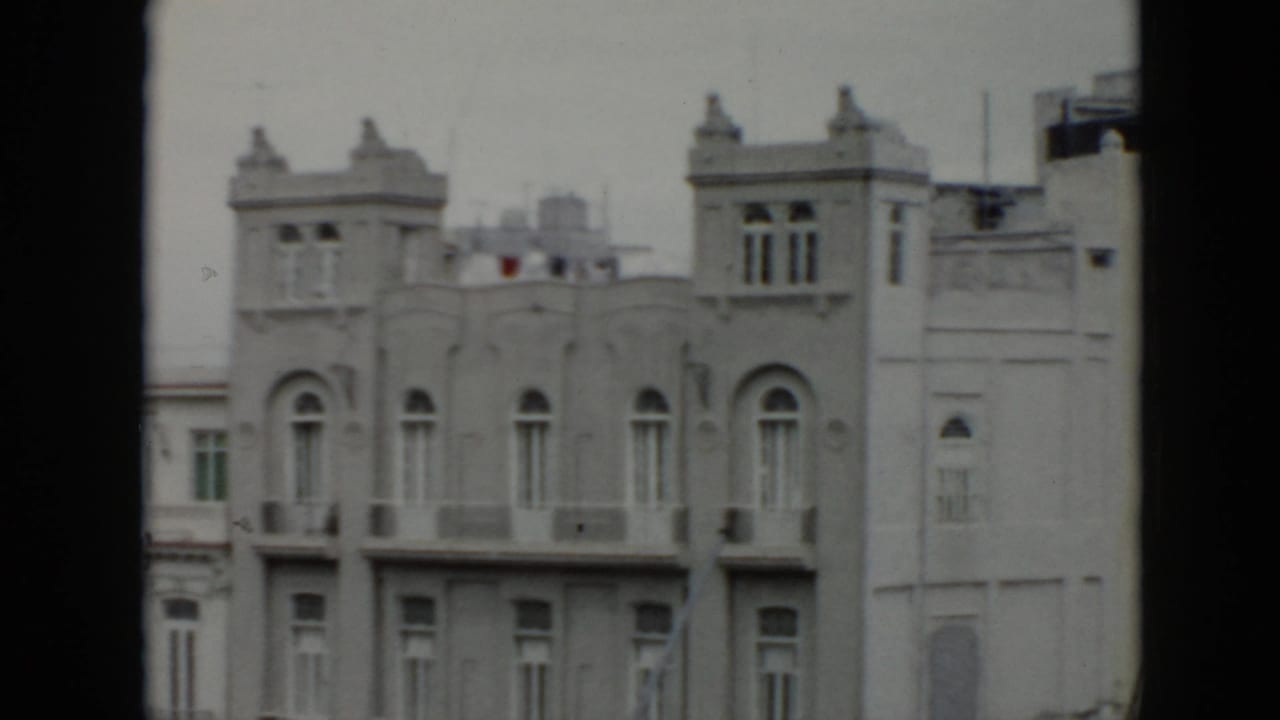 1956 Cuba home-movie frame showing a pale multi-story facade with arched windows, balconies, and decorative roofline towers.