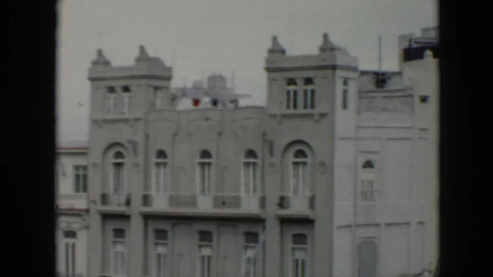 1956 Cuba home-movie frame showing a pale multi-story facade with arched windows, balconies, and decorative roofline towers.