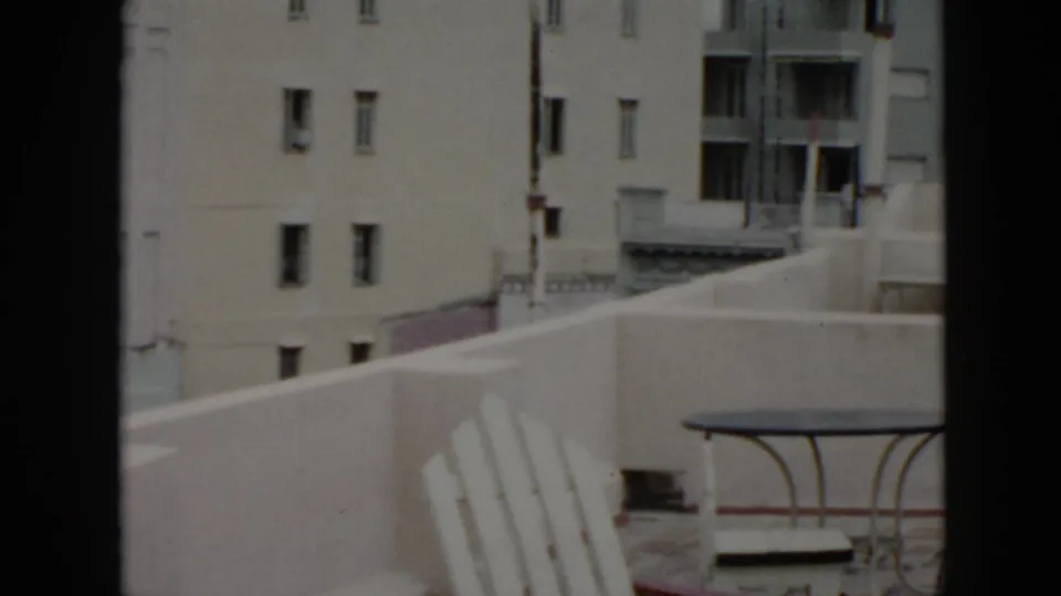 1956 Cuba home-movie frame showing balcony furniture in the foreground and pale multi-story buildings beyond.