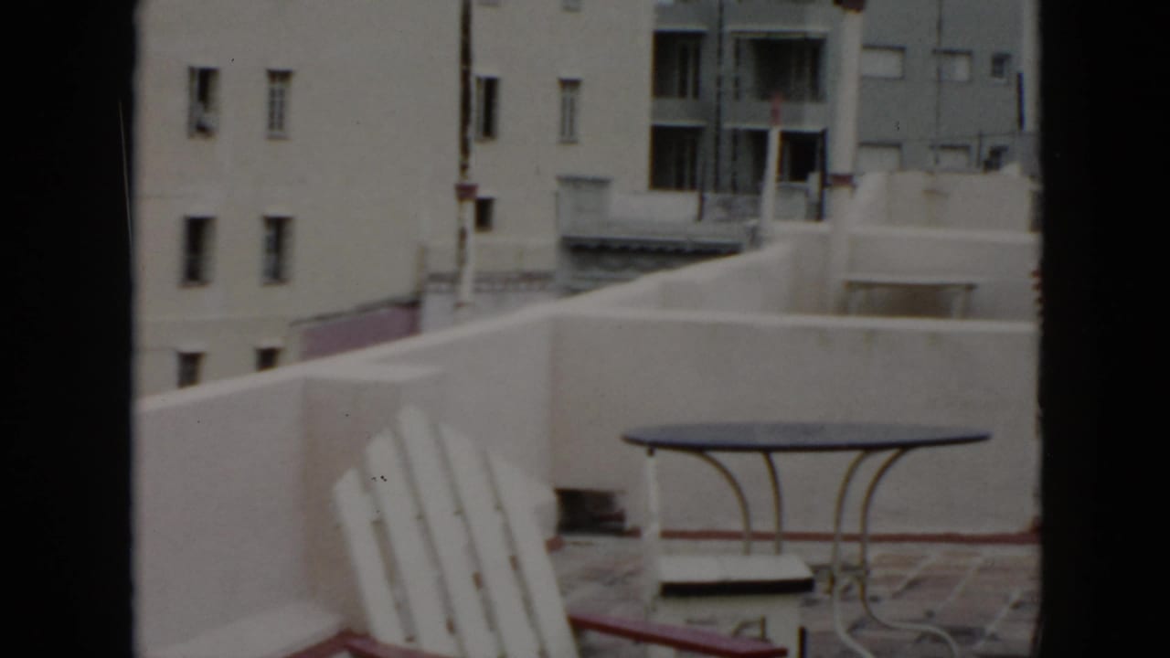 1956 Cuba home-movie frame showing a rooftop patio with white chairs, a small metal table, and neighboring apartment facades.