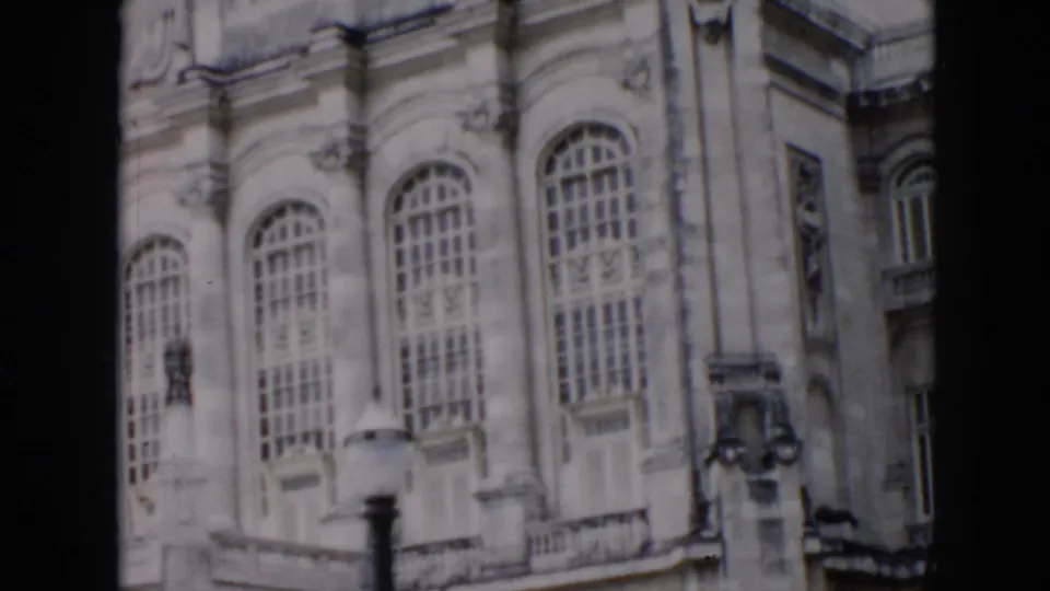 1956 Cuba 8mm frame of the ornate building facade with tall arched windows, balcony stonework, carved trim, and a dark foreground post.