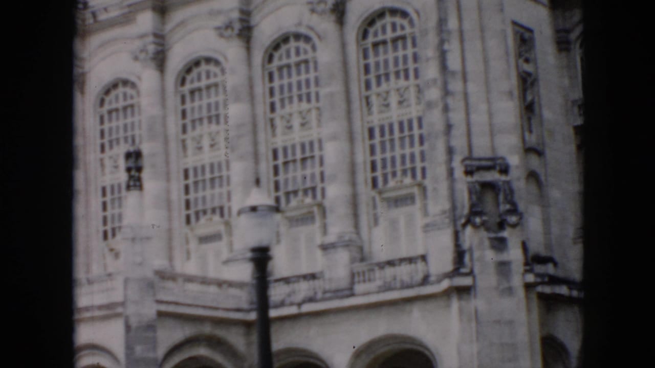 1956 Cuba home-movie frame of an ornate historic facade with arched windows, masonry details, and a streetlamp in the foreground.