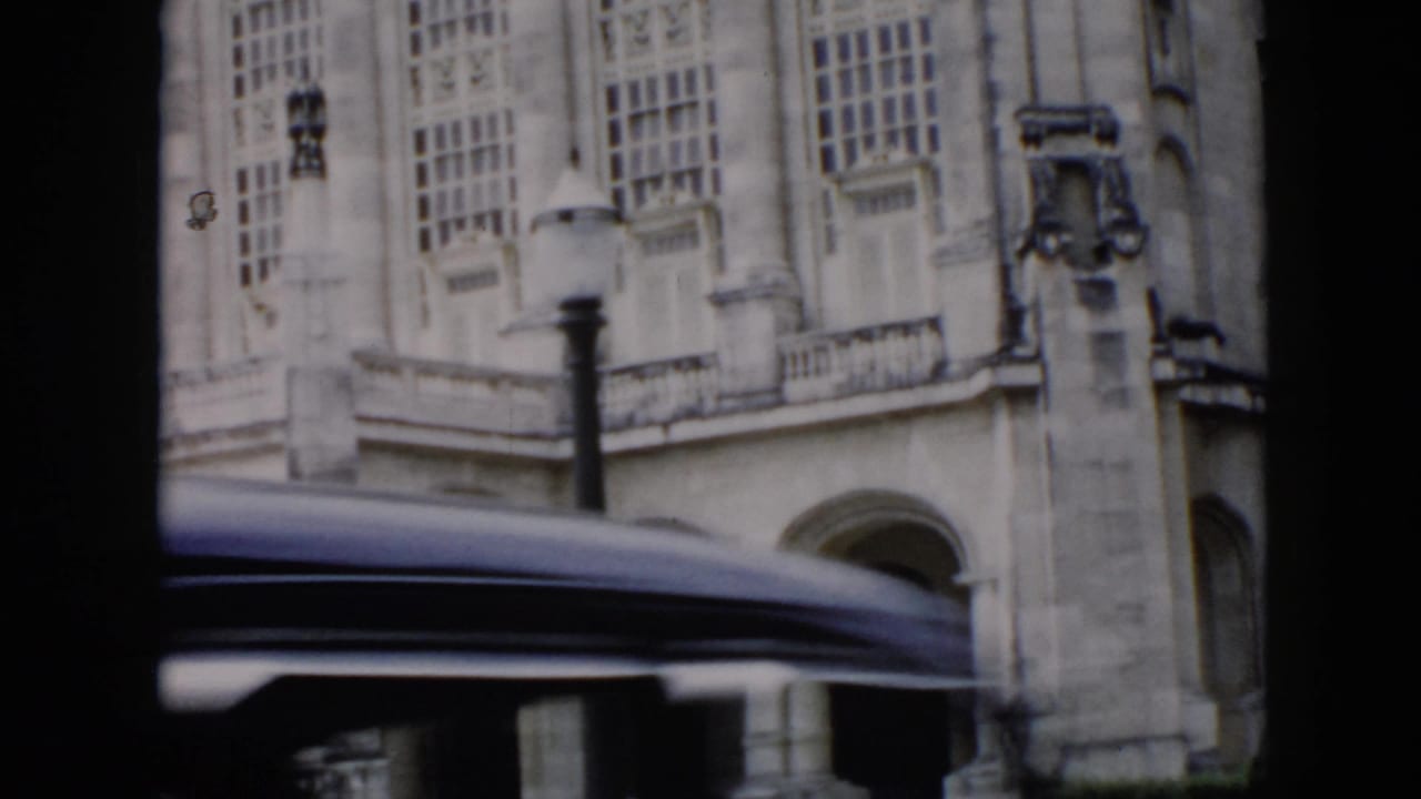 A 1956 Cuba driving shot frames the historic building through dark car silhouettes, with tall windows and a passing roofline across the view.