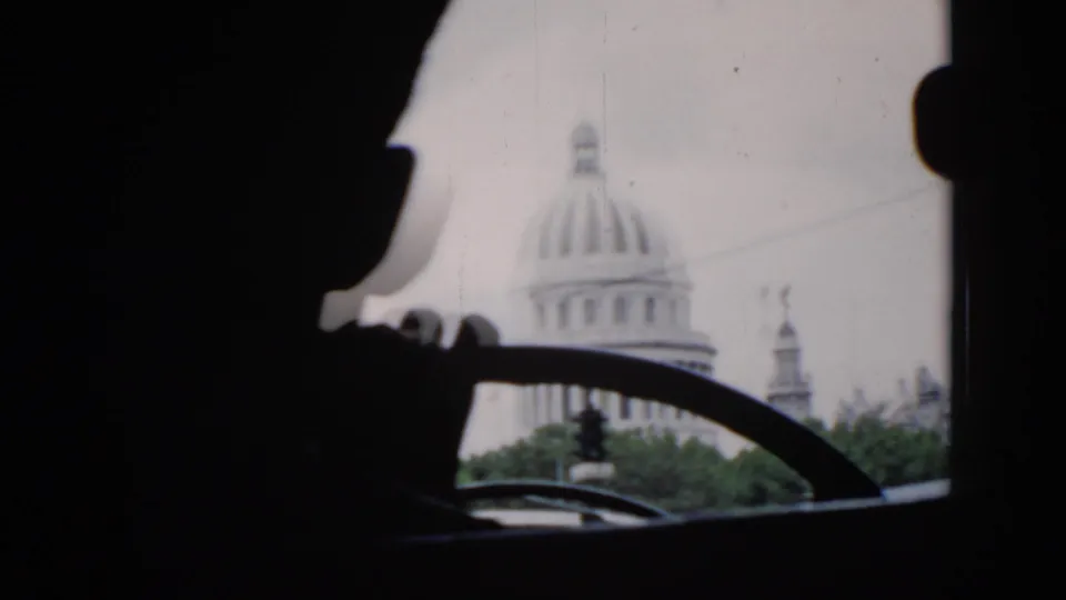 Inside a car in 1956 Cuba, a driver silhouette appears in the foreground while a domed landmark is visible beyond the windshield.