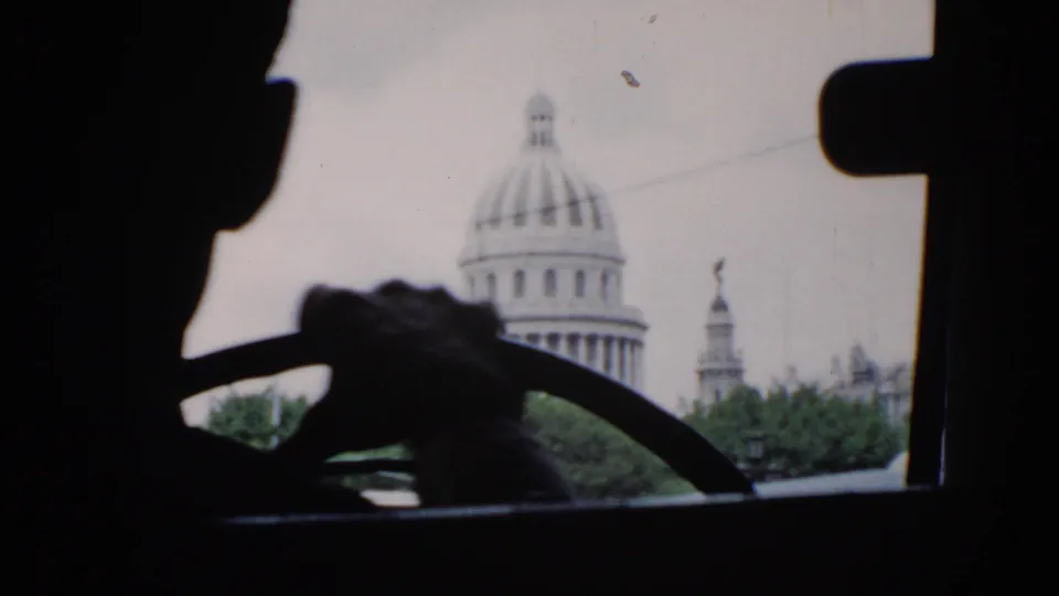 1956 Cuba home-movie frame from inside a car, with the driver's hands on the wheel and a domed capitol-style building beyond trees.