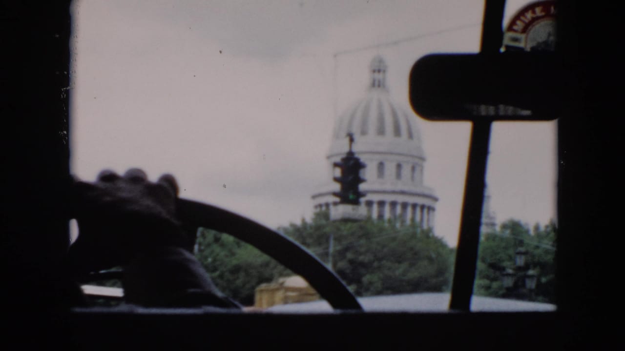 Passenger-side view from a moving car in Cuba, showing a domed capitol-style building, street signal, windshield posts, and mirror.