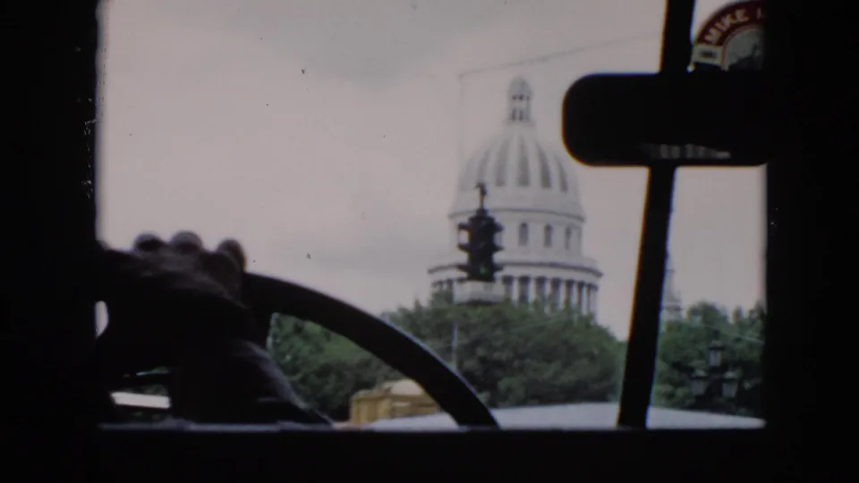 Passenger-side view from a moving car in Cuba, showing a domed capitol-style building, street signal, windshield posts, and mirror.