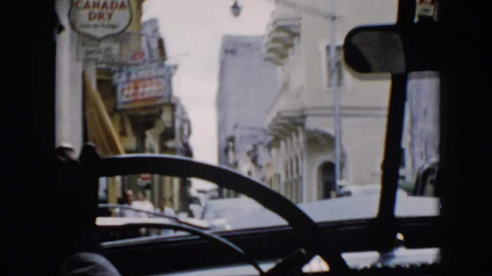1956 Cuba street view from inside a moving car, with storefront signage, pedestrians, parked or passing cars, and bright building facades.