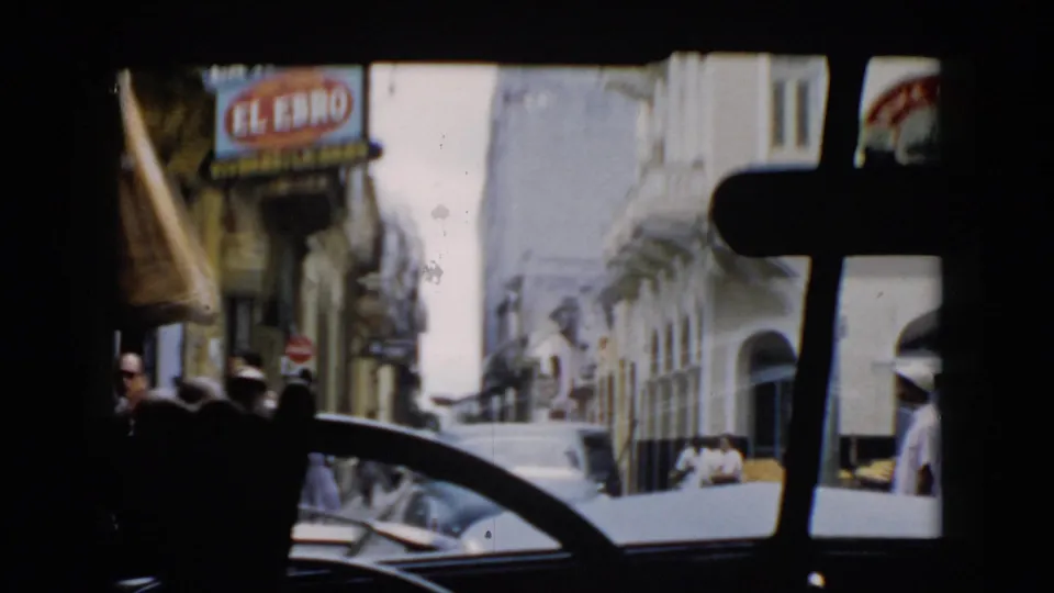 Cuba home-movie frame looking through a windshield at a busy street with an El Ebro storefront sign, pedestrians, cars, and pale building fronts.