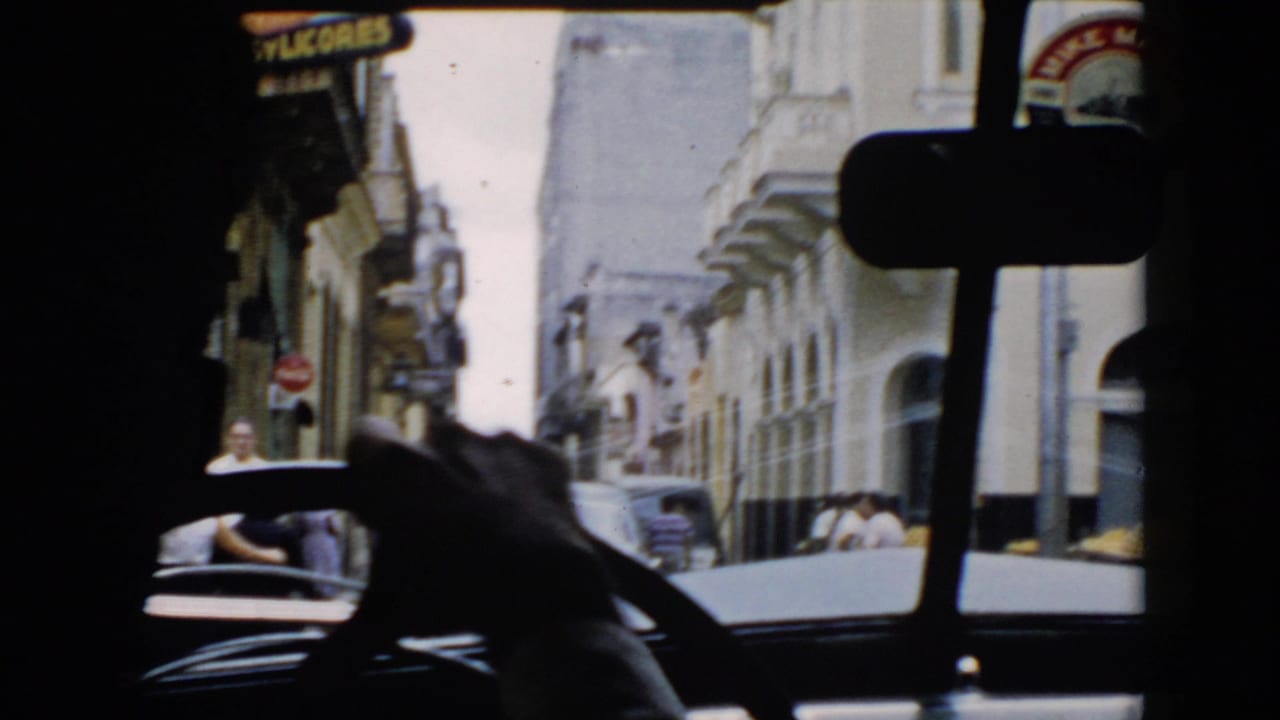 View through a car windshield onto a narrow street with storefronts, pedestrians, and Spanish signage.