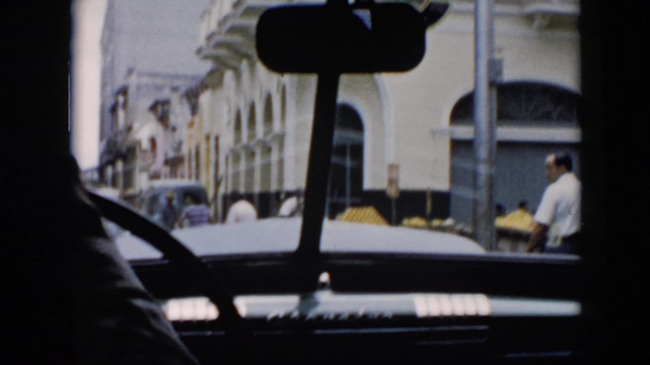 Interior car silhouette frames a street with arched buildings, pedestrians, and produce along the curb.