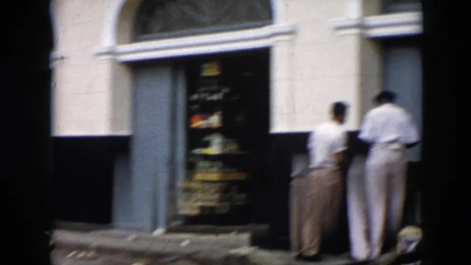 1956 Cuba street frame showing two men beside a storefront entrance and a curbside display that appears to include stacked oranges.