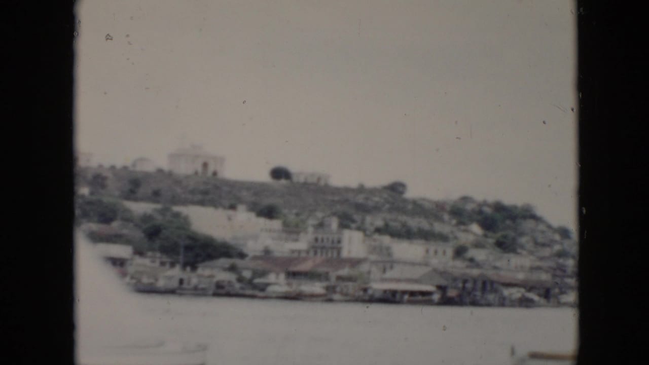 1956 Cuba home-movie frame showing waterfront structures, hillside buildings, and water seen through a dark vehicle window frame.