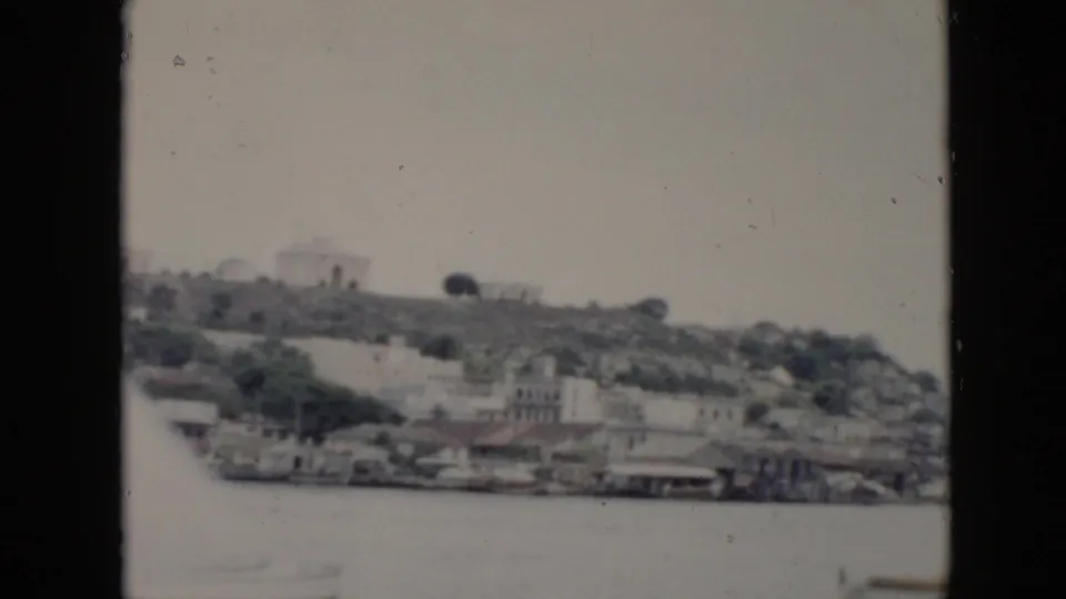 1956 Cuba home-movie frame showing waterfront structures, hillside buildings, and water seen through a dark vehicle window frame.