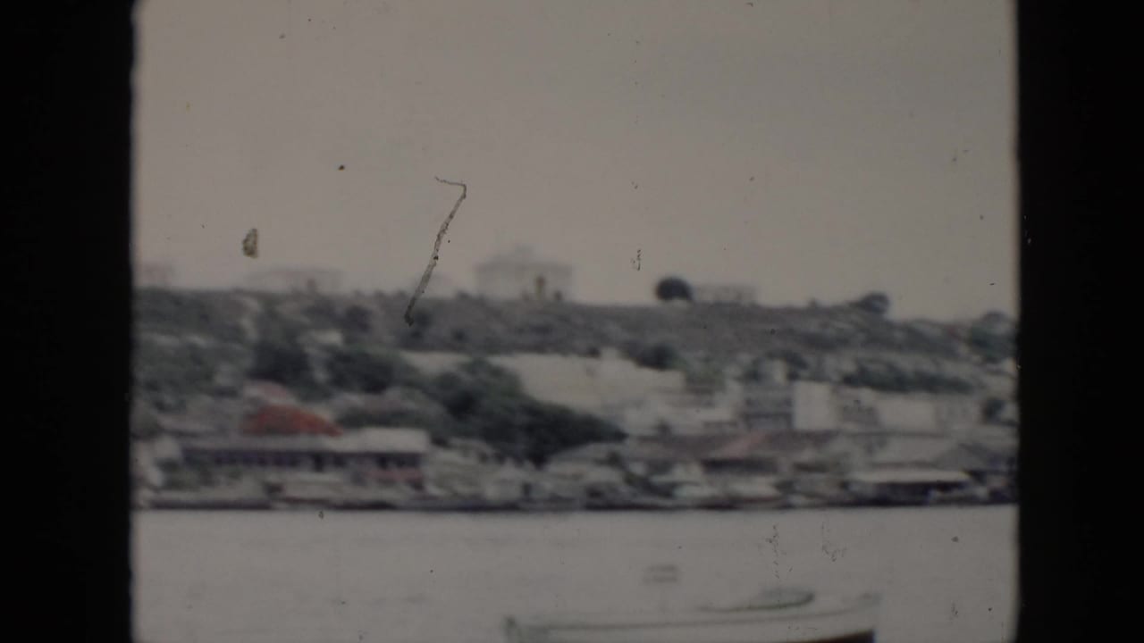 1956 Cuba travel frame with a distant shoreline, hillside structures, water, and visible 8mm film wear.