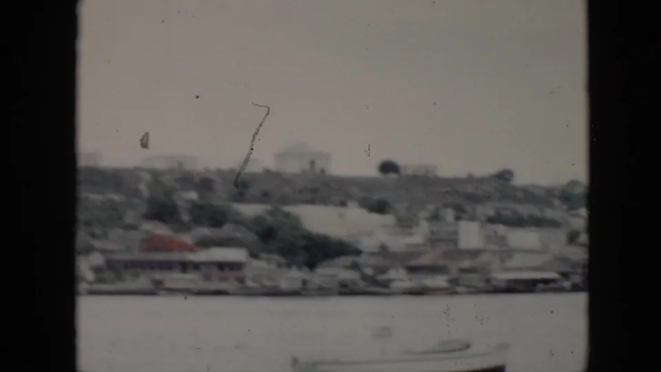 1956 Cuba travel frame with a distant shoreline, hillside structures, water, and visible 8mm film wear.