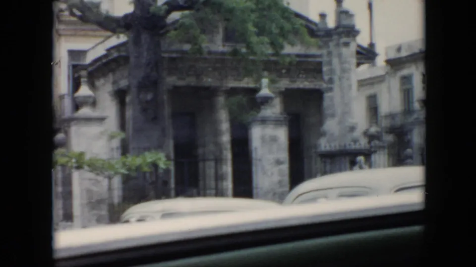 1956 Cuba street-level frame showing a masonry gate, tree, building facade, and white car roofline from a moving vehicle.